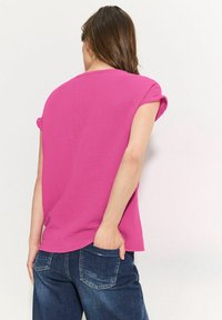 Donna con capelli lunghi e castani, che indossa una maglietta rosa a maniche corte e jeans blu scuro, rivolta di spalle con una mano nella tasca posteriore.