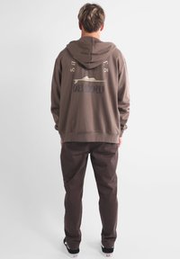 Felpa con cappuccio marrone con un design grafico che presenta palme e il nome del marchio sul retro, abbinata a pantaloni marroni coordinati.