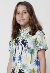 Camisa de manga corta con un patrón de palmeras en colores vibrantes de azul, verde, amarillo y negro sobre un fondo blanco, que cuenta con un cierre de botones en la parte delantera.