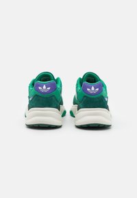 Un paio di sneaker Adidas verdi e viola viste di spalle, che mostrano le suole intermedie bianche e il logo a trifoglio Adidas sui talloni.