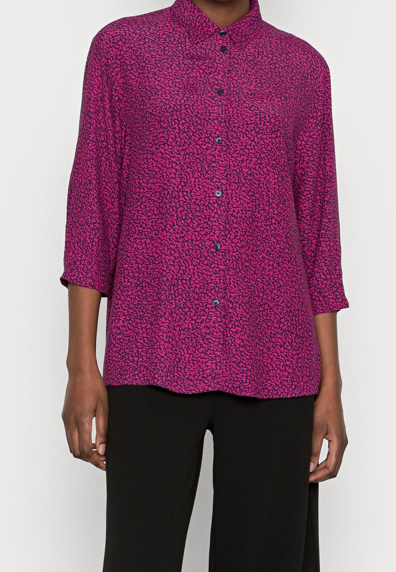 Blouse fuchsia avec un motif abstrait noir, à boutons sur le devant et manches trois-quarts, fabriquée en tissu léger.
