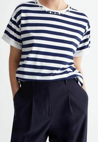 Camisa de rayas en azul marino y blanco con un escote redondo adornado con cuentas decorativas. Manga corta, corte holgado, combinada con pantalones oscuros.