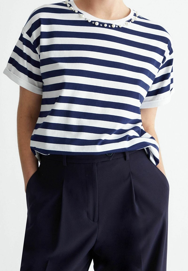 Camisa de rayas en azul marino y blanco con un escote redondo adornado con cuentas decorativas. Manga corta, corte holgado, combinada con pantalones oscuros.