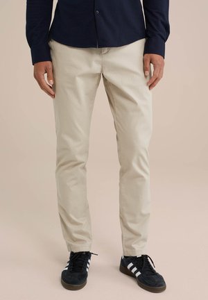 Mann trägt beige Chinos, schwarz-weiße Sneaker und ein dunkelblaues Langarmshirt vor einem einfarbigen Hintergrund.