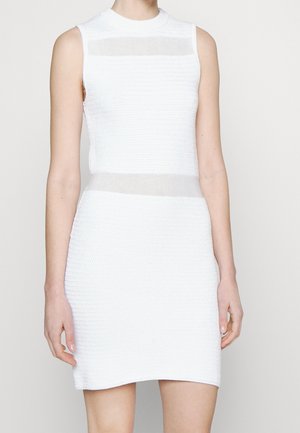 Robe en maille blanche sans manches avec des panneaux horizontaux transparents au niveau de l'encolure et de la taille portée par une personne debout sur un fond uni.