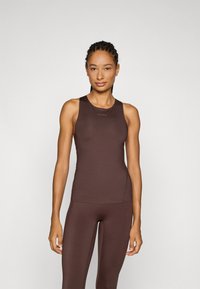 Vrouw met een gevlochten haar, gekleed in een strakke bruine mouwloze top en bijpassende leggings, staande tegen een neutrale achtergrond.