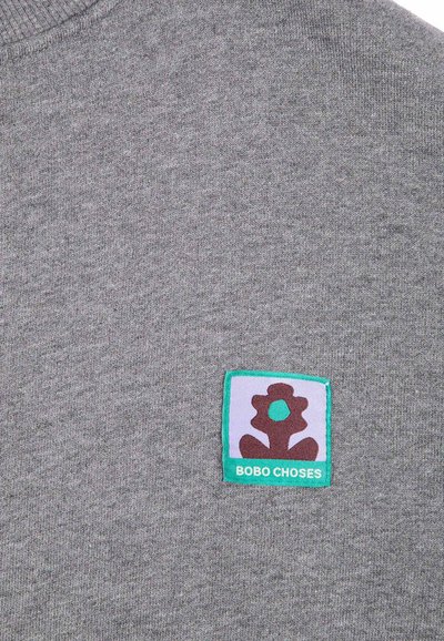 Tissu gris avec un petit patch carré montrant une fleur brune stylisée avec un centre turquoise et le texte "BOBO CHOSES" en turquoise.
