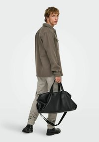 Sac de voyage en cuir noir avec deux poignées supérieures, bandoulière ajustable et finition lisse. Le modèle porte un haut en polaire gris et un pantalon clair.