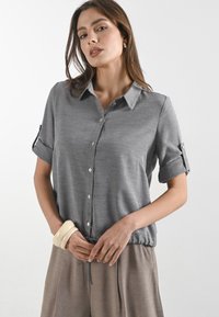 Blusa grigia a maniche corte con colletto, chiusura a bottoni e maniche arrotolate. Il tessuto è liscio con una vestibilità comoda, abbinata a pantaloni color sabbia.