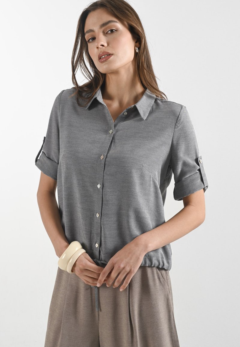 Blusa grigia a maniche corte con colletto, chiusura a bottoni e maniche arrotolate. Il tessuto è liscio con una vestibilità comoda, abbinata a pantaloni color sabbia.