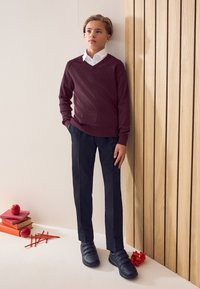 Bordó kötött V-nyakú pulóver, fehér galléros ing, fekete szabott nadrág és fekete tépőzáras sportcipő, fa borítású fal előtt.