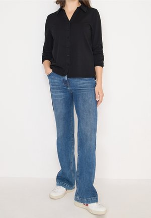 Femme portant une chemise noire boutonnée, un jean bleu droit avec des bandes latérales, des baskets blanches, debout avec une main dans la poche.
