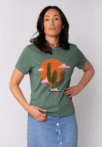 Groen katoenen T-shirt met een cactusprint tegen een oranje zon, met lichtroze wolken erboven. Korte mouwen, ronde hals.