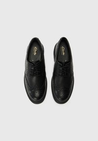 Sorte skinnlær sko med brogue-design, perforeringer, rund tå og snøring, satt mot en ensfarget bakgrunn.