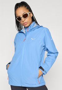 Veste imperméable bleu clair avec fermeture éclair devant, capuche à cordon, accents contrastés et poignets réglables. Convient pour les activités de plein air.