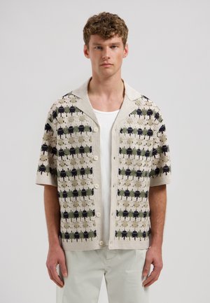 Jonge man die een crèmekleurige gebreide korte mouw cardigan draagt met een zwart-groen geruit patroon over een wit overhemd en lichte broek.