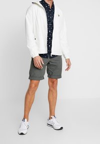 Uomo in giacca bianca a zip sopra una camicia a fantasia blu scuro, pantaloni corti grigi arrotolati e sneaker bianche, in piedi su uno sfondo semplice.