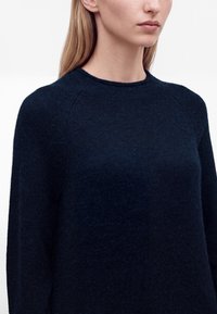 Jeune femme portant un pull uni bleu marine foncé à col rond avec manches longues, visible des épaules aux lèvres sur fond blanc.