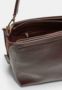 Sac à main en cuir brun avec une forme structurée. Il présente des éléments en métal doré, deux compartiments zippés et une courte bandoulière.