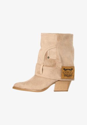 Beige suède midkuitlaars met gestapelde houten hak, spitse neus, riemdetail en een bruine leren patch met zwarte afbeelding aan de buitenkant.