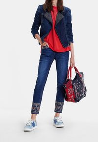 Kurtka dżinsowa z fakturowanymi rękawami, czerwona koszula, haftowane dżinsy, niebieskie i białe tenisówki oraz torba materiałowa z wzorem w czerwone akcenty.