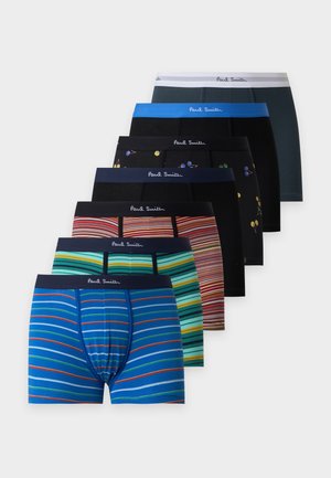 Homme musclé debout, pieds nus, portant un boxer bleu foncé avec une ceinture blanche sur fond blanc uni.