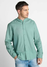 Man in een lichtgroen overhemd met mandarinekraag en blauwe jeans, die naar rechts kijkt met één hand in zijn zak.