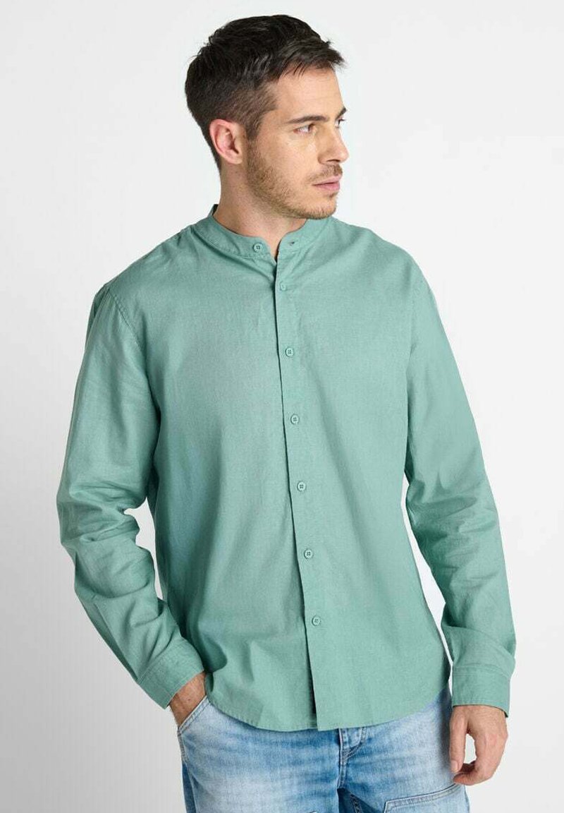 Man in een lichtgroen overhemd met mandarinekraag en blauwe jeans, die naar rechts kijkt met één hand in zijn zak.