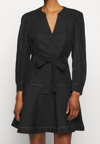 Mustassa mekossa on V-pääntie, pitkät hihat ja vyötä ympäröivä solmittava vyö. Mekossa on valkoista ompelua helman ja hihojen varrella. A-linjainen siluetti.