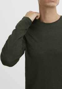 Chemise à manches longues vert foncé avec un col rond. Le tissu semble doux avec une fine texture. Les manches ajustées atteignent les poignets.