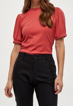 Punainen ribbineulottu paita, jossa on lyhyet, liehukkeet hihoissa ja pyöreä pääntie, yhdistetty mustiin räätälöityihin housuihin, joissa on sivutaskut ja nappikiinnitys.