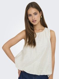 Blouse sans manches en crème clair avec un motif texturé subtil, décolleté rond et coupe décontractée, associée à un jean en denim bleu foncé.