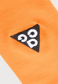 Μαύρο τριγωνικό ένθετο με λευκά κεντημένα γράμματα "gpp" σε πορτοκαλί υφή φόντου από ύφασμα.