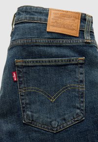 Πίσω τσέπη σκούρων μπλε τζιν Levi's με κίτρινη ραφή, κόκκινη ετικέτα Levi's και καφέ δερμάτινη ετικέτα με το λογότυπο στη ζώνη της μέσης.