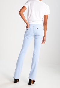 Light blue flared jeans with a smooth texture and a mid-rise fit, featuring back pockets and a small logo patch. Paired with a white T-shirt.