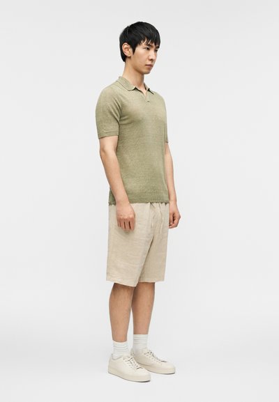 Polo manches courtes vert olive, shorts en lin beige, baskets crème et chaussettes blanches, debout contre un fond blanc uni.