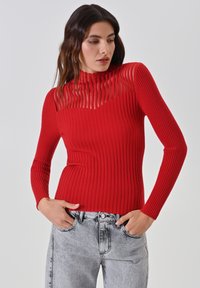 Roter gerippter Pullover mit hohem Kragen und transparenter gestreifter Einlage über der Brust. Enganliegendes Design mit langen Ärmeln. Getragen mit grauen, ausgebeulten Jeans.