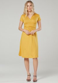 Gelbes knielanges Kleid mit kurzen Ärmeln, Wickeldesign und gerafftem Taillenbereich, aus einem weichen Stoff. Kombiniert mit schwarzen Riemchensandalen.