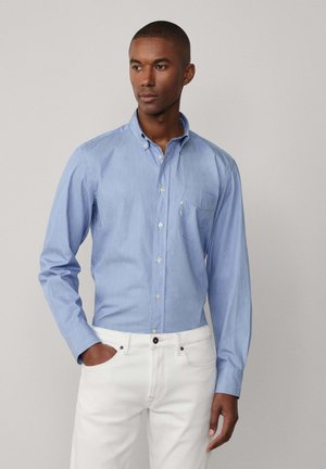 Himmelblaues Hemd mit langen Ärmeln, einem Kragen und einer Brusttasche; aus weichem Stoff; kombiniert mit weißen Hosen.