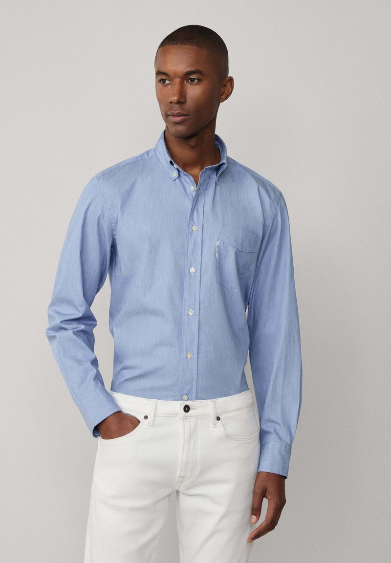 Camicia azzurra chiara con maniche lunghe, colletto e taschino sul petto; realizzata in tessuto morbido; abbinata a pantaloni bianchi.