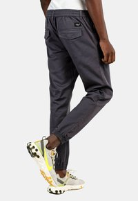 Pantalones jogger de color gris oscuro con cintura elástica, bolsillos en los laterales y en la parte posterior, tobillos acampanados y textura suave. Llevados con zapatillas gris claro con acentos.