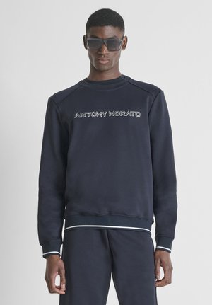 Man draagt donkerblauwe sweatshirt en broek, zonnebril, met "ANTONY MORATO" logo op de voorkant van de sweatshirt, staand tegen een effen achtergrond.