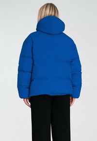 Niebieska kurtka puchowa z dużym kapturem, luźny krój i pikowana faktura. Posiada prosty dół i długie rękawy, w zestawie z czarnymi spodniami.