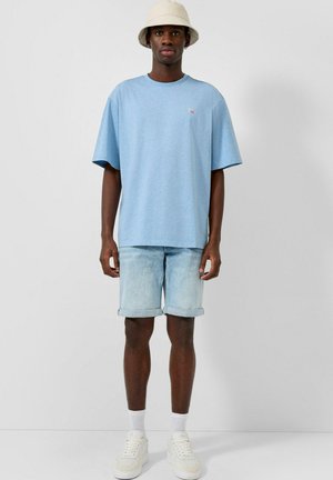 Mężczyzna stoi, ubrany w jasnoniebieską oversize koszulkę, jasne krótkie spodenki dżinsowe, białe skarpetki, białe sneakersy i beżowy kapelusz typu bucket na jednolitym tle.