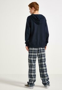 Eine rothaarige Person trägt einen dunklen Hoodie, marineblaue und weiße karierten Hosen und braune Converse-Sneaker. Sie steht mit dem Rücken zu einer einfarbigen Hintergrund.