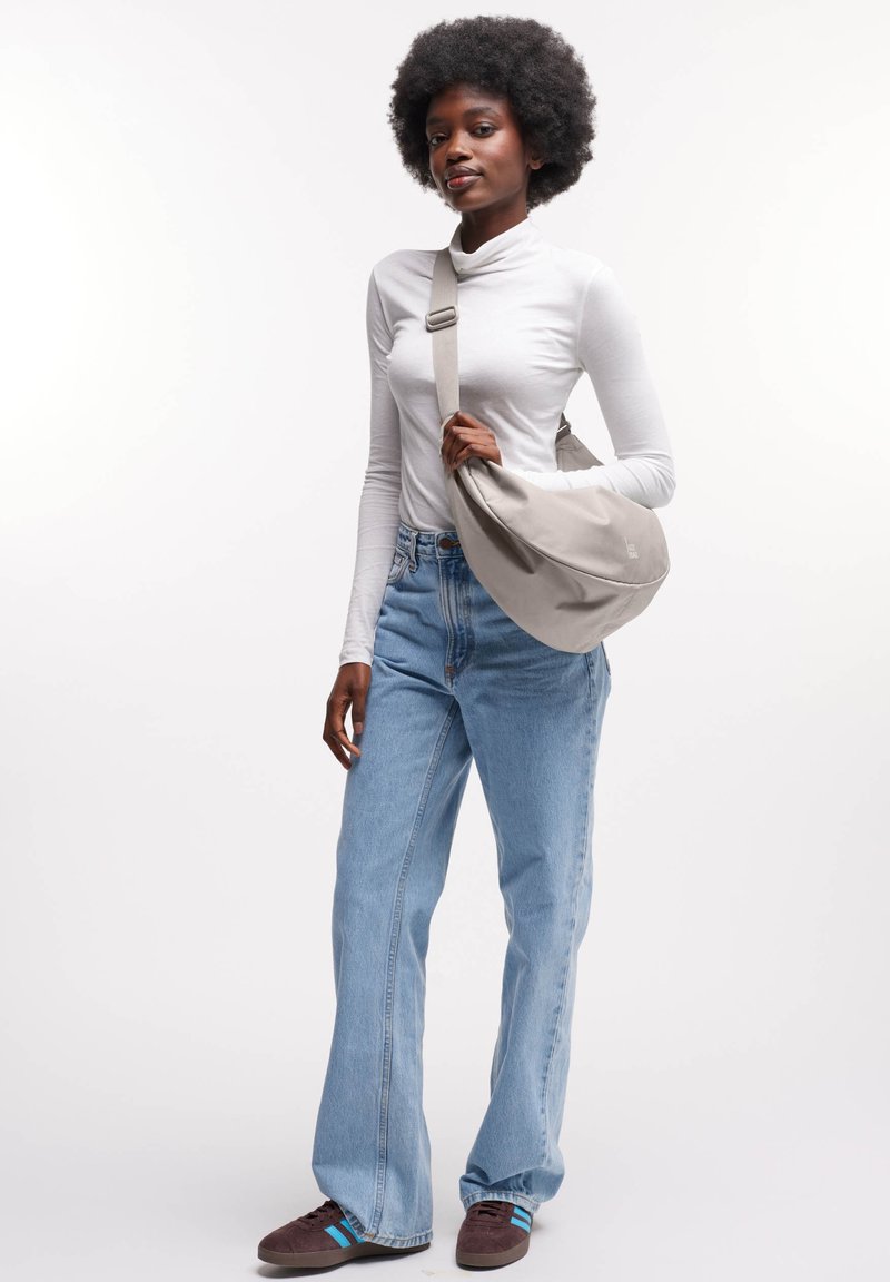 Grijze schoudertas van zacht stof, met schouderriem, kromme vorm. Model draagt een wit longsleeve shirt, high-waisted lichtblauwe spijkerbroek en bruine sneakers.