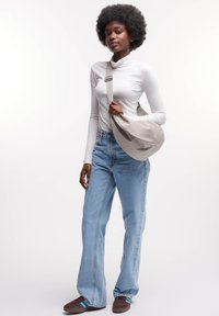 Borsa a tracolla grigia realizzata in morbido tessuto, con tracolla e forma curva. Modello indossa una maglietta bianca a maniche lunghe, jeans chiari a vita alta e sneakers marroni.