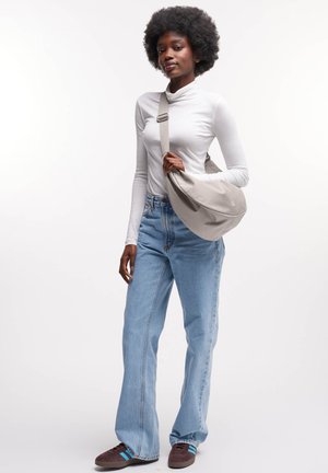Borsa a tracolla grigia realizzata in morbido tessuto, con tracolla e forma curva. Modello indossa una maglietta bianca a maniche lunghe, jeans chiari a vita alta e sneakers marroni.