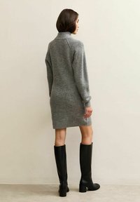 Abito grigio lavorato a maglia con colletto alto, texture a coste e maniche lunghe, abbinato a stivali neri con tacco al ginocchio su uno sfondo neutro.