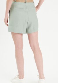 Hellgr�üne Shorts mit glatter Textur, ausgestattet mit Gesäßtaschen und einem lockeren Schnitt, kombiniert mit weißen Turnschuhen.